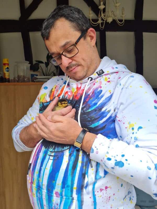 Man in colourful hoodie gently cradling a duckling against his chest, looking down with care, in a traditional British home.